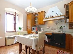 A kitchen with wooden cabinets, a dining table set for two, a built-in oven, and a window with blinds. - Apartment 1872 (Fazana)