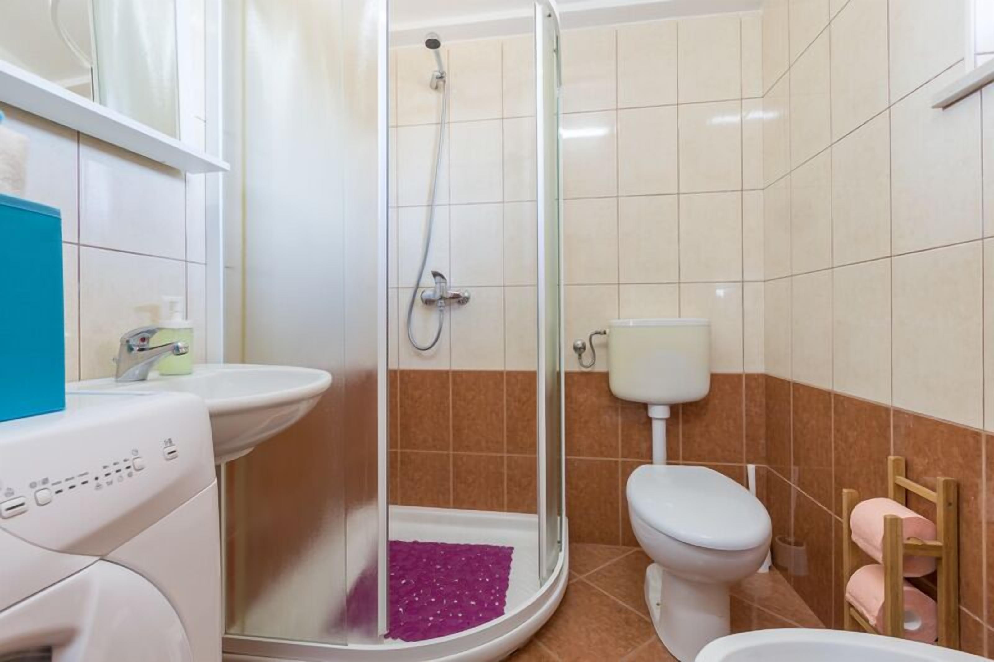 A bathroom with a glass shower enclosure, a white toilet, a sink, and a washing machine.