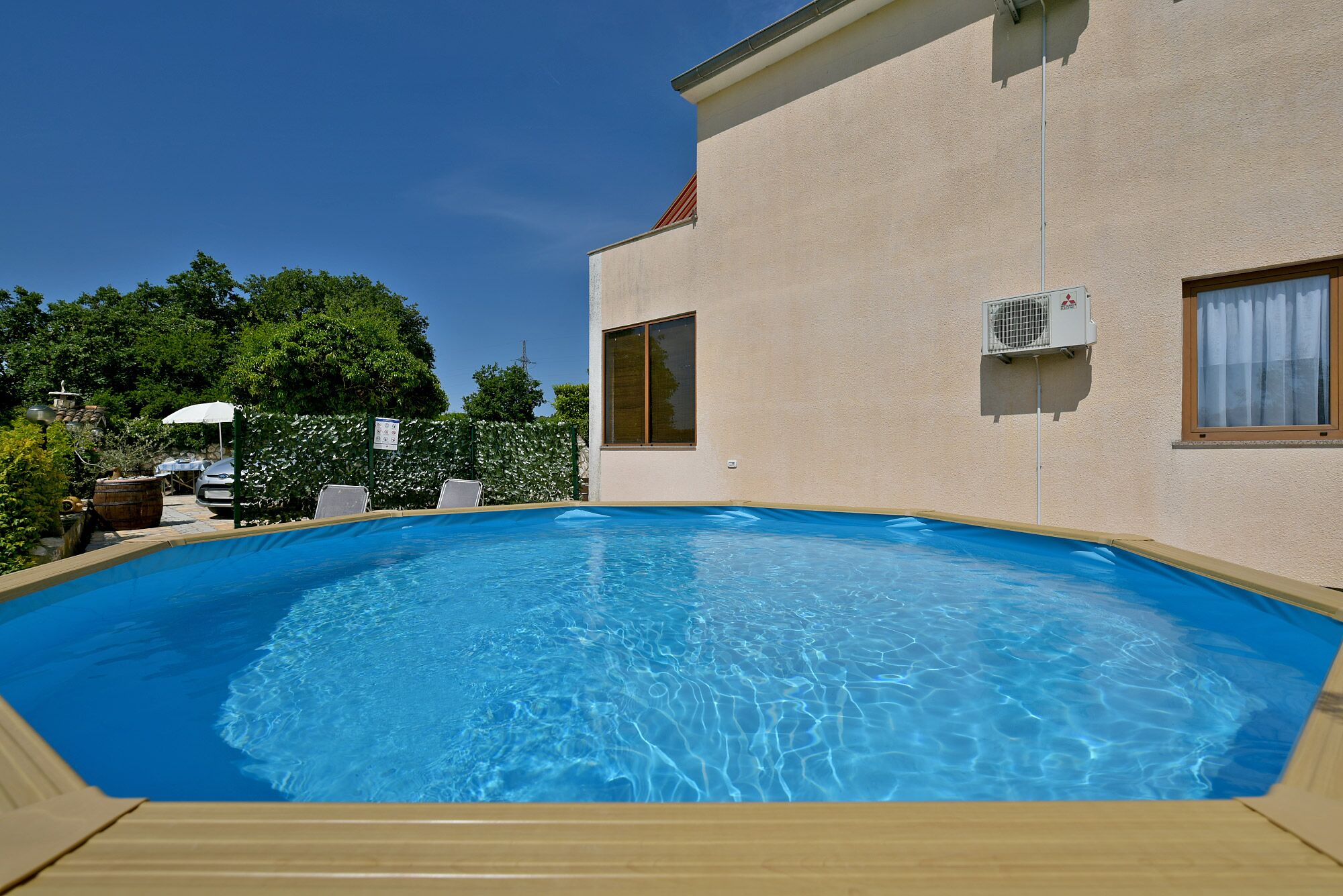 A swimming pool with a wooden deck, surrounded by a beige building with two windows and an air conditioning unit.