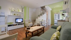 Un soggiorno moderno con un tavolino in legno, un divano verde e un televisore che mostra un programma. Si notano anche sedie bianche, una scala e un angolo cucina con pensili.