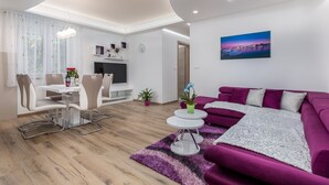 Una sala de estar moderna con un sofá púrpura, un comedor con una mesa y sillas blancas, un televisor y un cuadro en la pared.