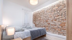 Un dormitorio con pared de piedra, cama con ropa de cama gris, mesita de noche con lámpara y ventana con cortinas ligeras.