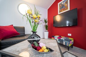 Una sala de estar moderna con una pared de acento roja, un televisor de pantalla plana sobre un otoman colorido, un florero con flores sobre una mesa de centro de mármol y un sofá gris con un cojín rojo.