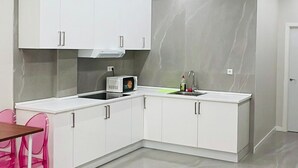 A modern kitchen with white cabinets, a microwave, and a sink. There is a pink chair and a table. - Hostel 18 (Madrid)