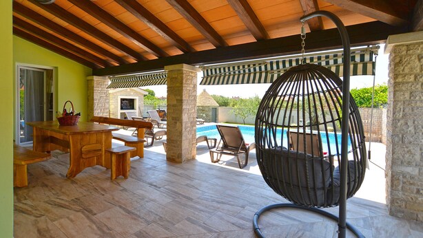 Un espacio exterior techado con piscina, muebles de madera y una hamaca colgante.