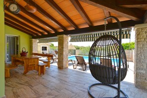 A covered outdoor area with a pool, wooden furniture, and a hanging chair.