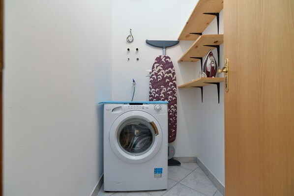 A compact laundry room with a front-loading washing machine, a drying rack, and a wooden shelf with a clock and decorative items.