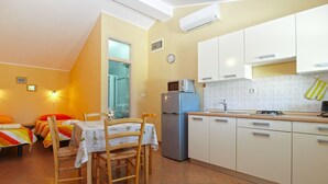 A compact kitchen with white cabinets, a refrigerator, a microwave, and a sink. There is a dining table with chairs and a small window. - Apartment 1895 (Marcana)