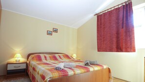 A neatly made bed with a plaid bedspread, two bedside lamps, a small drawer, and a window with a red curtain. - Apartment 1895 (Marcana)