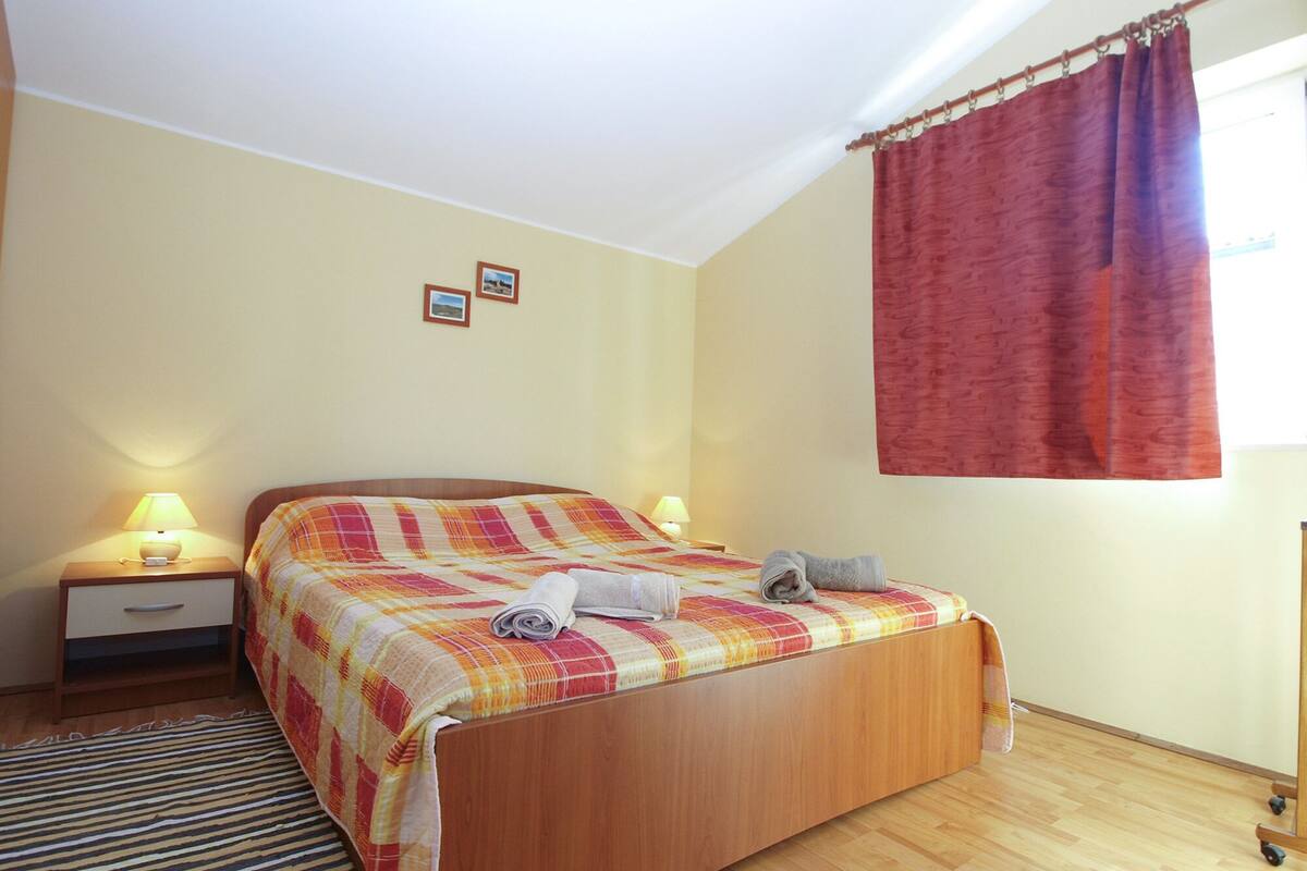 Una cama bien hecha con una colcha de cuadros, dos lámparas de noche, un cajón pequeño y una ventana con cortina roja.