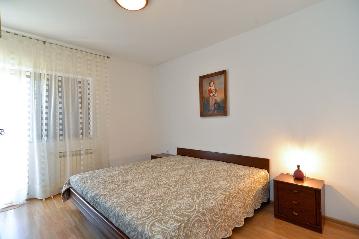 A bedroom with a bed, a nightstand, a lamp, a painting on the wall, and a window with sheer curtains.
