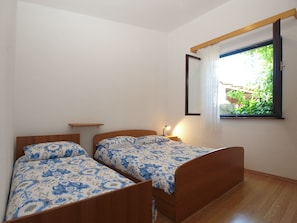 A bedroom with two wooden beds, a nightstand with a lamp, a small shelf, and a window with a view of greenery. - Apartment 1907 (Zminj)