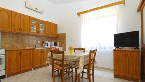 A compact kitchen with wooden cabinets, a dining table with chairs, a TV on a stand, and a window with curtains. - Apartment 1907 (Zminj)