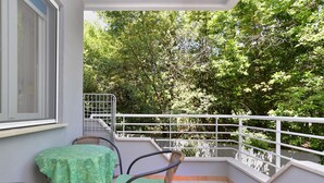Ein Balkon mit einem grünen Tisch und Stühlen, einem weißen Geländer und Blick auf dichtes Blätterwerk.