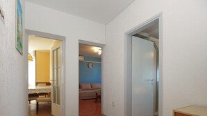 Um corredor com piso de madeira que leva a uma sala de jantar com mesa e cadeiras, uma sala de estar com sofá e um relógio na parede, e uma porta fechada.
