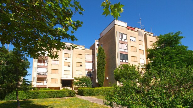 Um edifício residencial com vários andares e varandas, cercado por vegetação bem cuidada e um céu azul límpido.