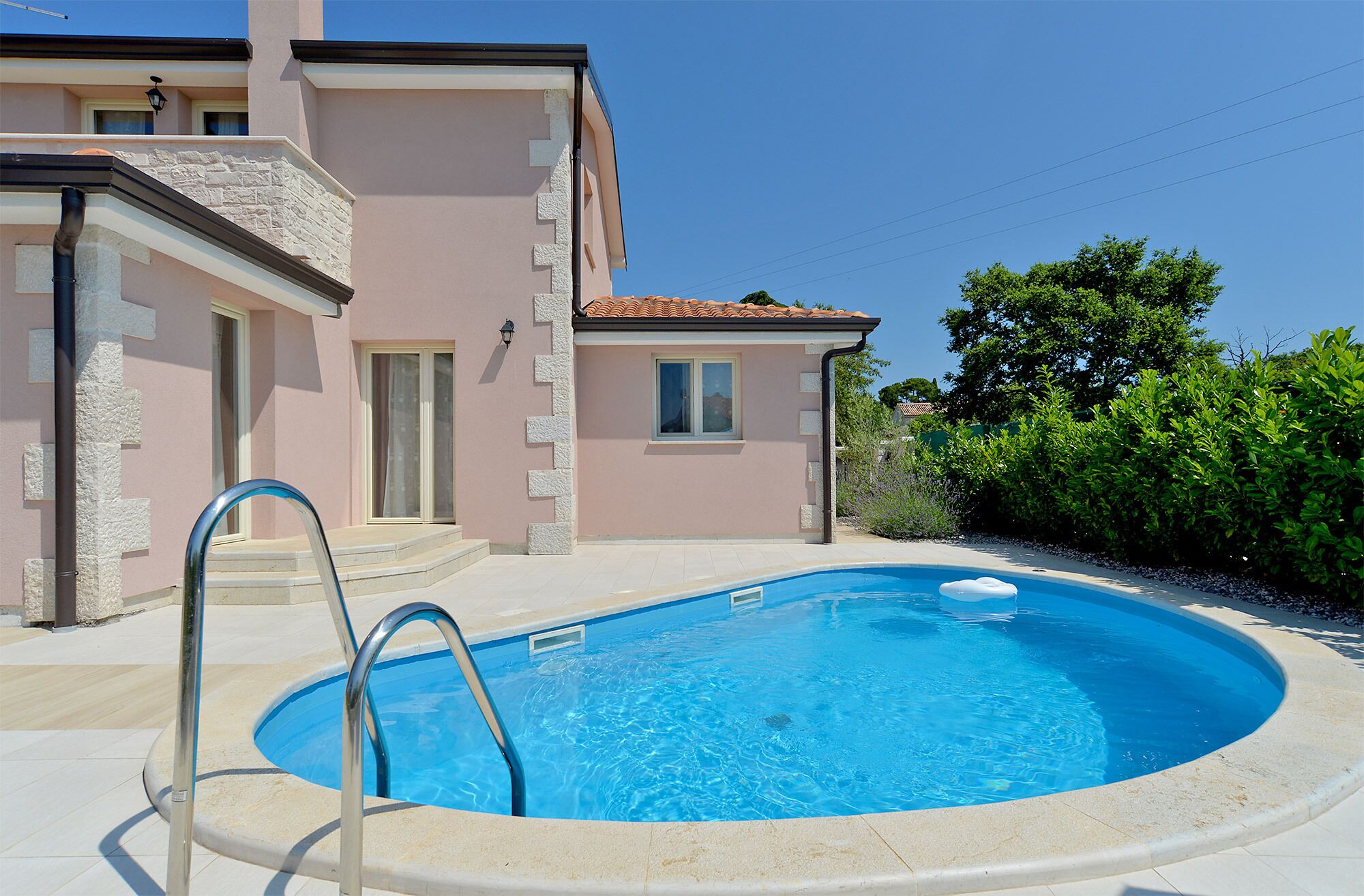 A swimming pool with a metal ladder, a house with a tiled roof, and a clear blue sky.