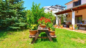 Jardí amb taula de fusta i plantes en testos, casa amb una pèrgola, i cel blau clar.