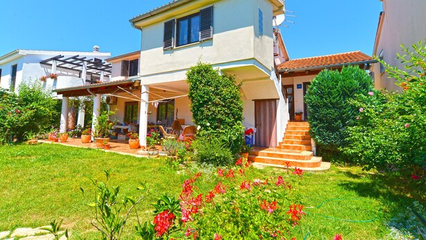 Una casa de dos pisos amb un porxo cobert, un jardí petit amb plantes en testos i una escala que porta a l'entrada.