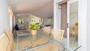 A spacious living area with a glass-top table surrounded by wicker chairs, a wooden door, and a view of a bedroom with a bed and a window. - House 1926 (Liznjan)
