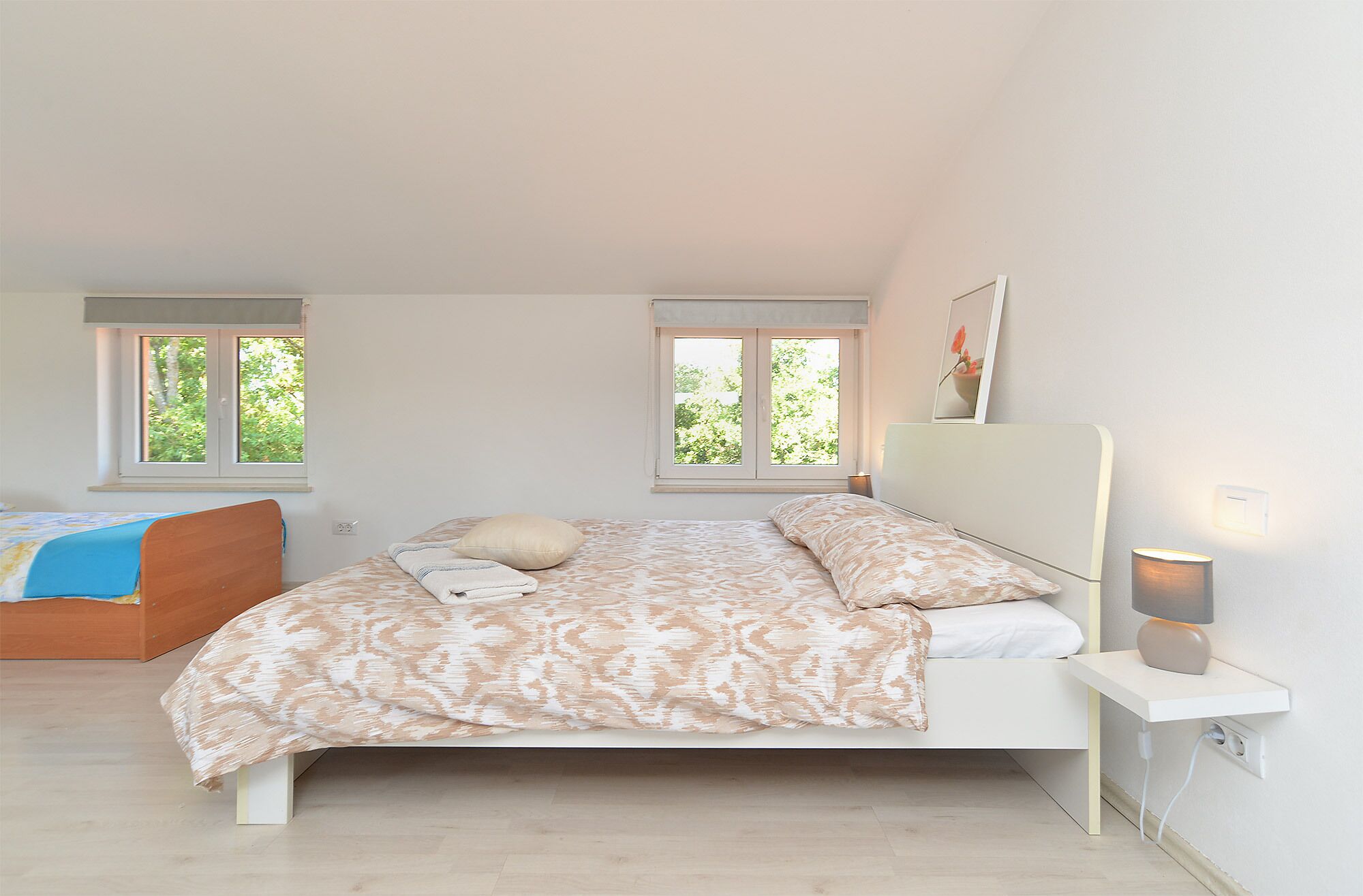 A bedroom with a bed, bedside table, lamp, and two windows with a view of greenery.