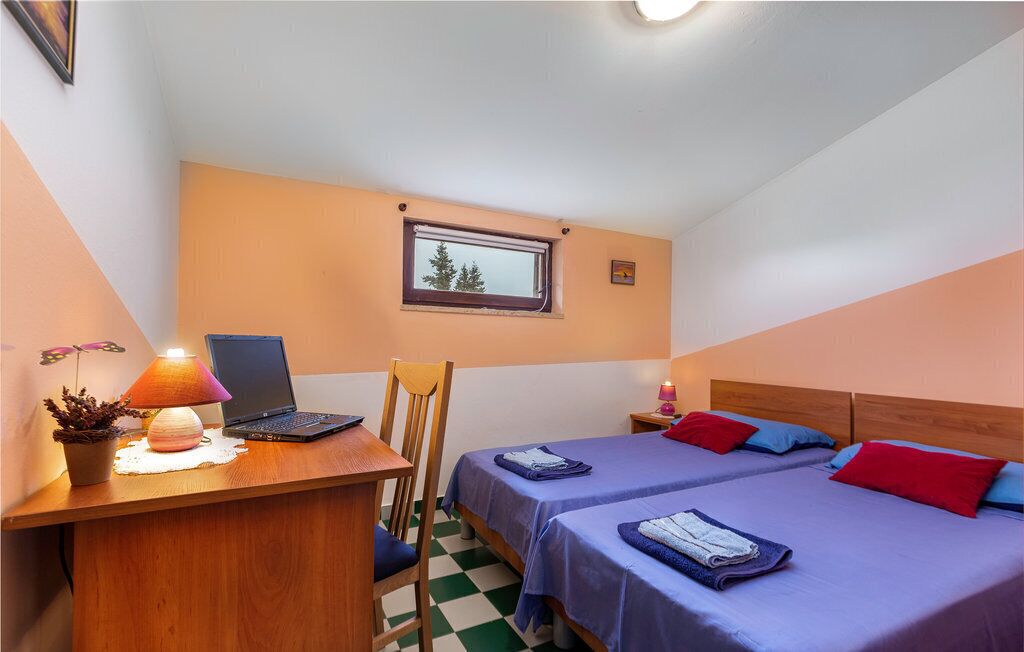A small, cozy room with two beds, a desk with a laptop, a chair, a window with a view of trees, and a lamp.