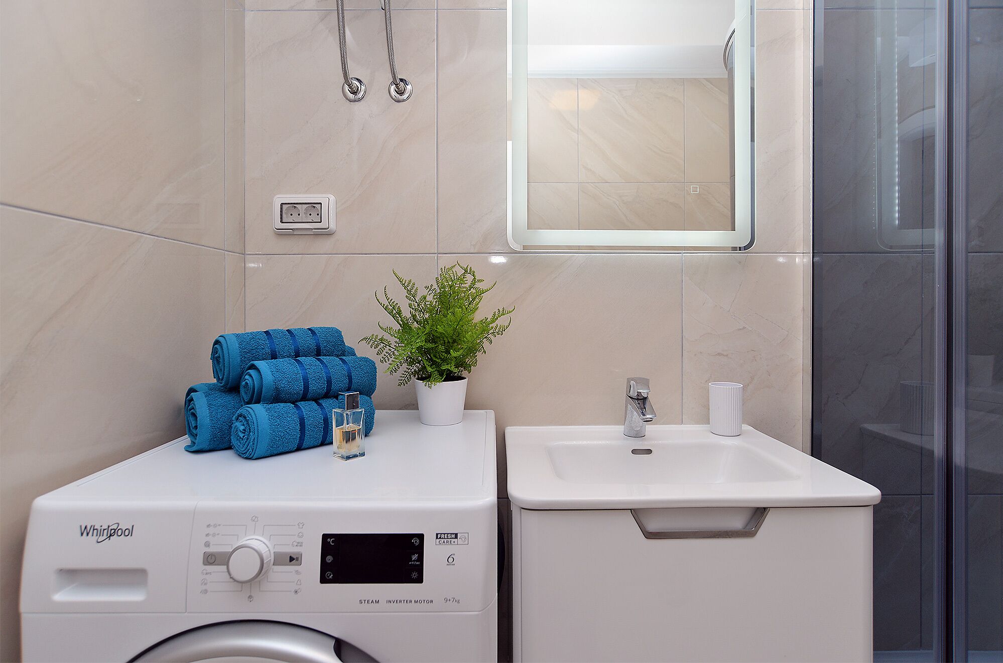 Casa de banho compacta com máquina de lavar e secar Whirlpool, uma pequena pia e uma planta em vaso.