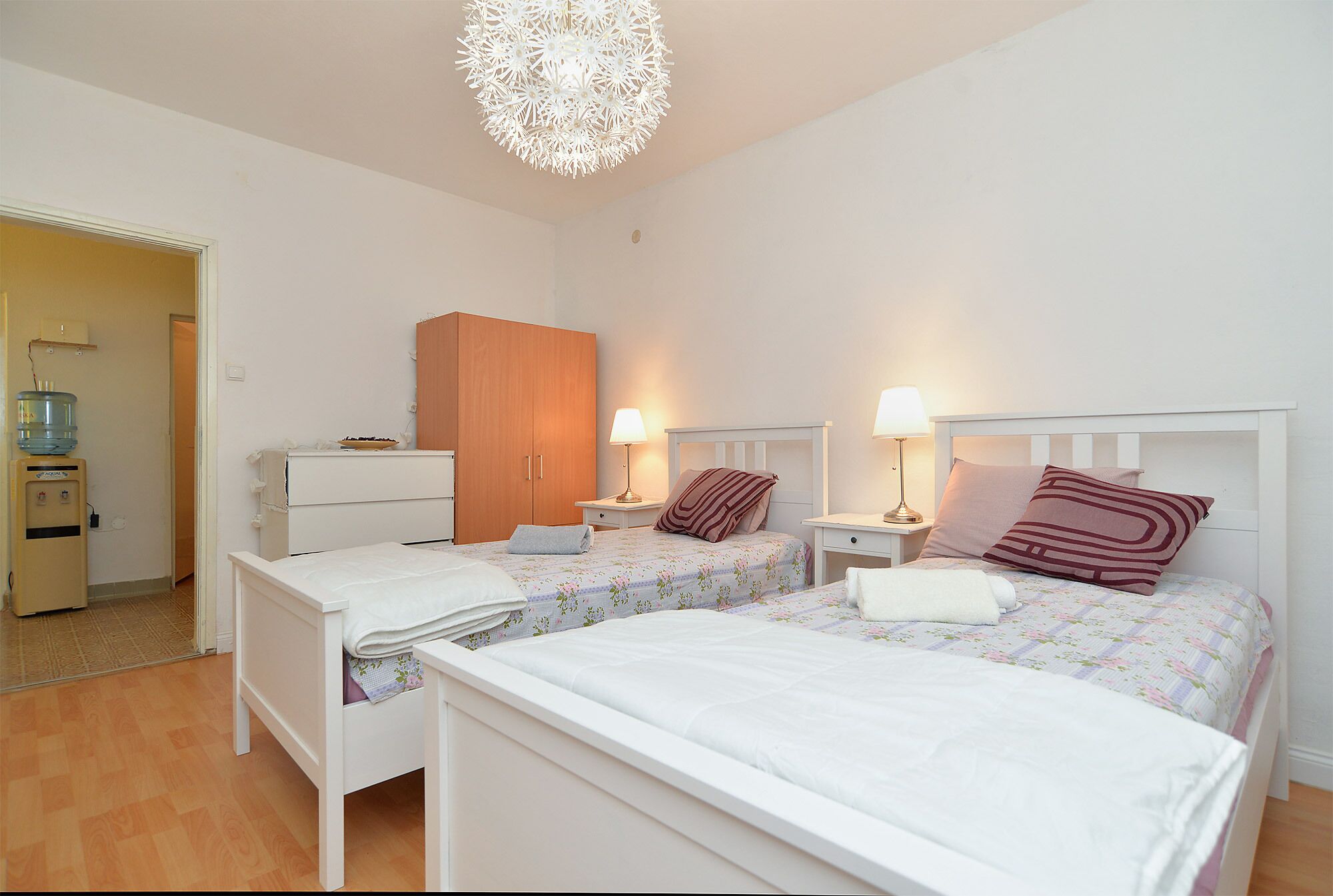 A bedroom with two beds, a wardrobe, bedside tables with lamps, and a water dispenser.