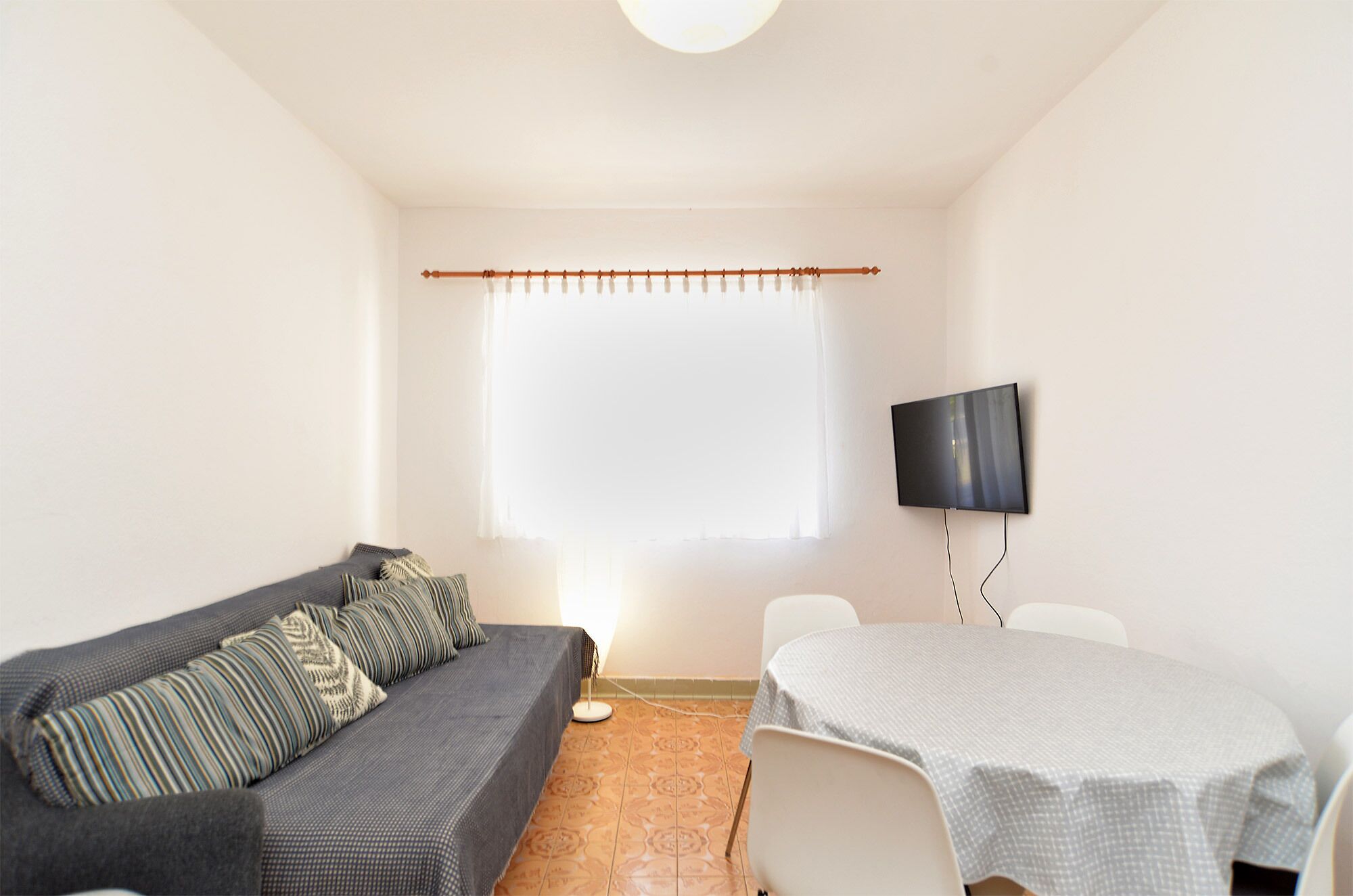 Een kleine, goed verlichte kamer met een grijze bank, een witte tafel en een flatscreen televisie die aan de muur is bevestigd.