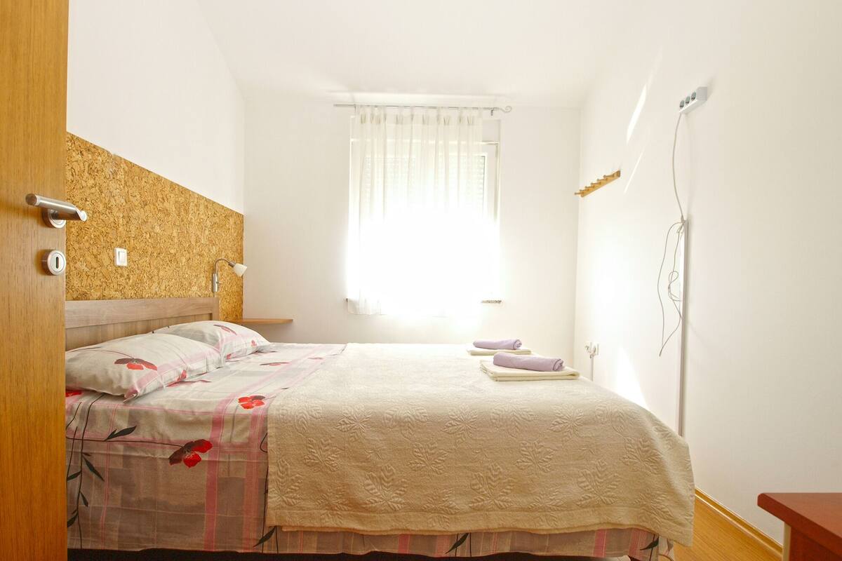A bedroom with a bed, a nightstand, a lamp, a window with curtains, and a wooden headboard.