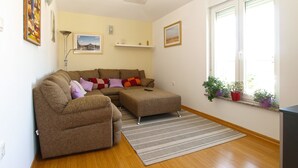 A living room with a brown sofa, a striped rug, a standing lamp, and a window with plants. - Apartment 1932 (Fazana)