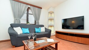 Sala de estar com um sofá cinzento, duas almofadas azuis, uma mesa de centro de vidro com copos de vinho e uma travessa de petiscos, uma televisão de tela plana e um móvel de entretenimento de madeira.
