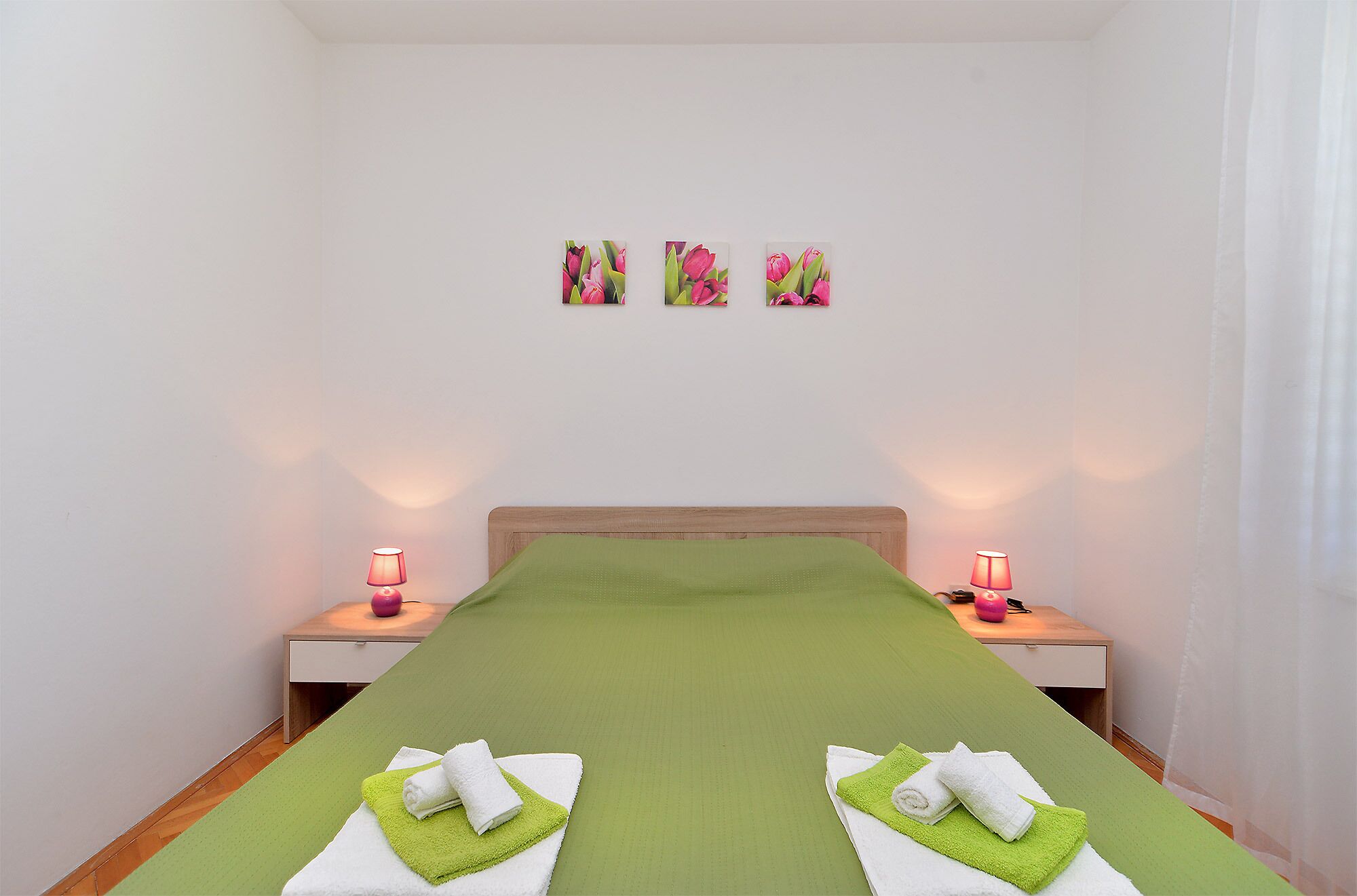 A neatly made bed with green bedding and two towels on green mats, two bedside lamps, and three wall-mounted pictures of pink flowers.