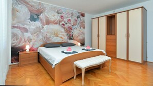 Quarto com um grande mural floral, uma cama de madeira, um banco, uma mesa de cabeceira com abajur e um guarda-roupa.
