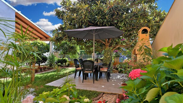 Um pátio externo com mesa e cadeiras, cercado por vegetação exuberante e plantas floridas.