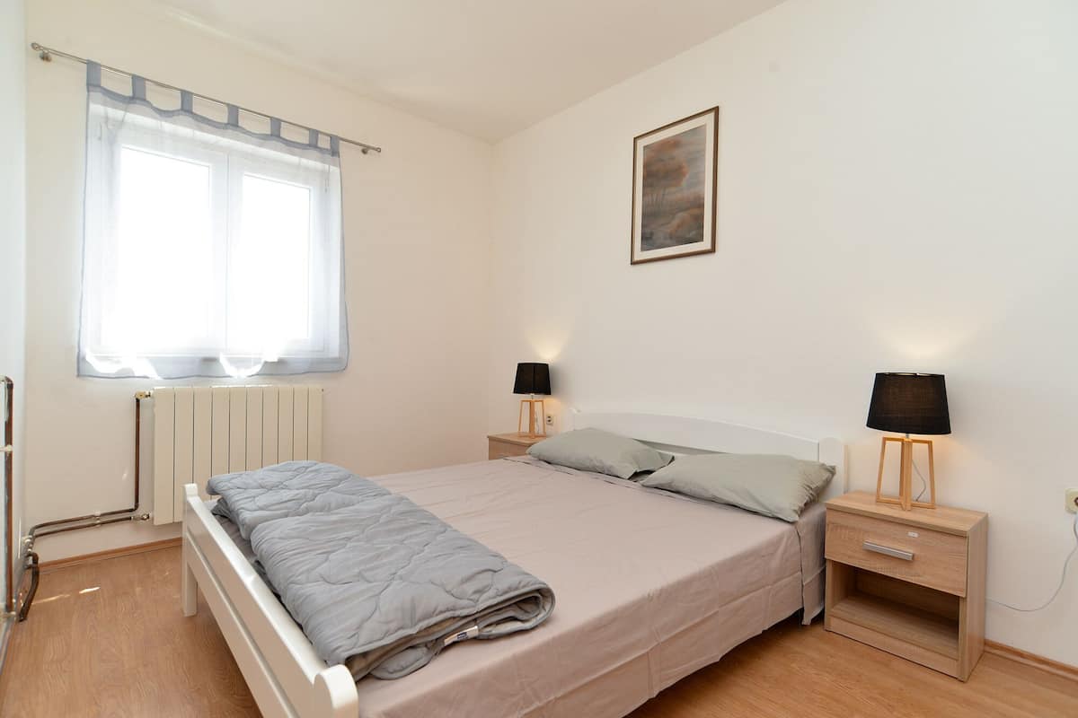 Une chambre à coucher avec un lit, deux tables de chevet avec des lampes, un radiateur, une fenêtre avec des rideaux et un tableau encadré au mur.