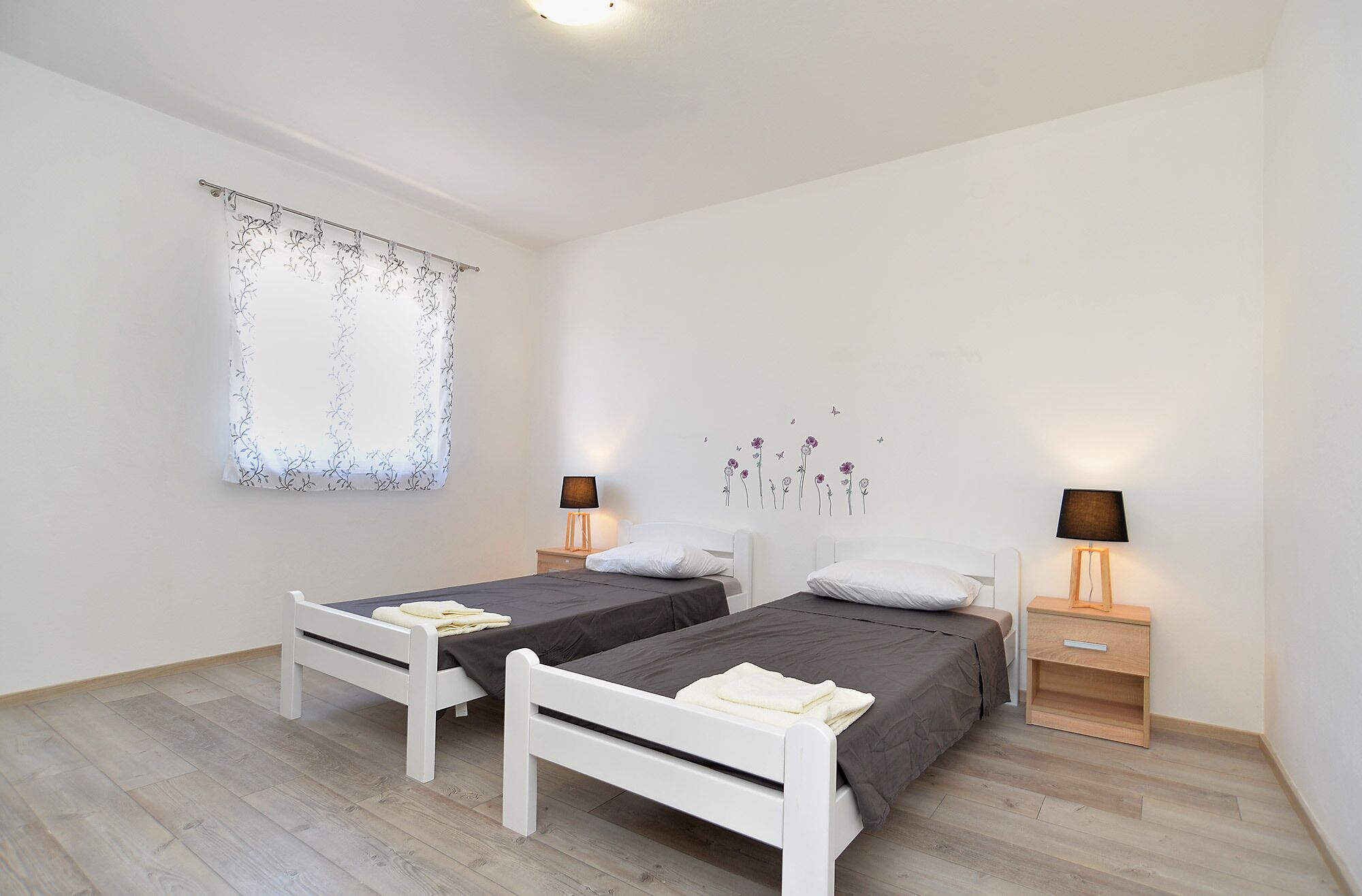 A bedroom with two single beds, a window with a floral curtain, a wall decal of flowers and birds, and bedside lamps.