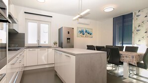 A modern kitchen with a central island, a dining area with a table and chairs, and a large window with blinds. - House 1939 (Pula)