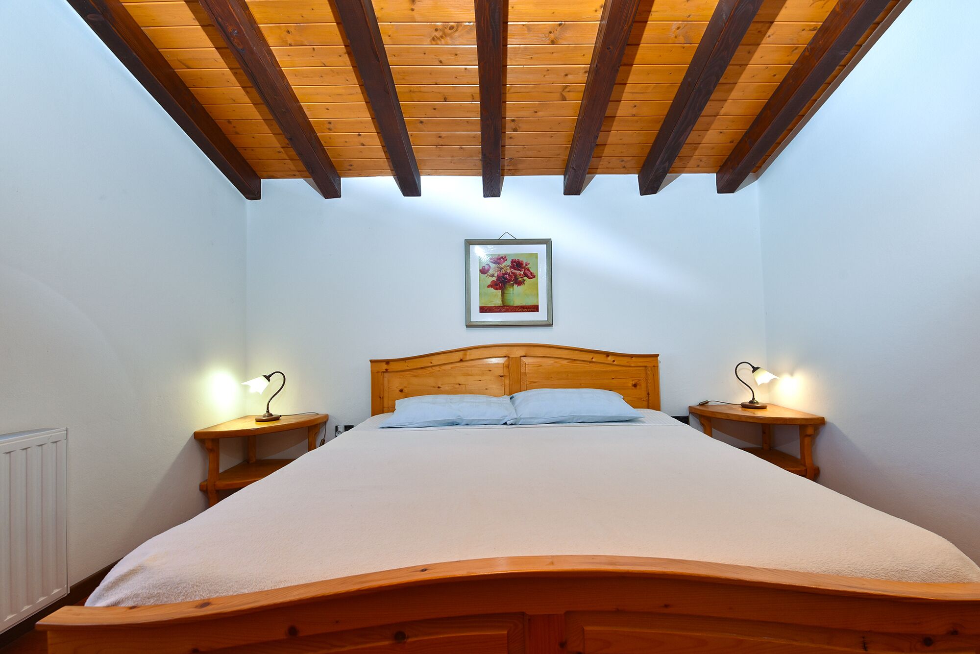 A bedroom with a wooden bed, two bedside tables with lamps, a wooden ceiling, and a framed picture on the wall.