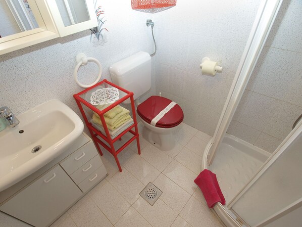 Un baño con lavamanos, inodoro y ducha. Hay un taburete rojo con toallas y un adorno encima.