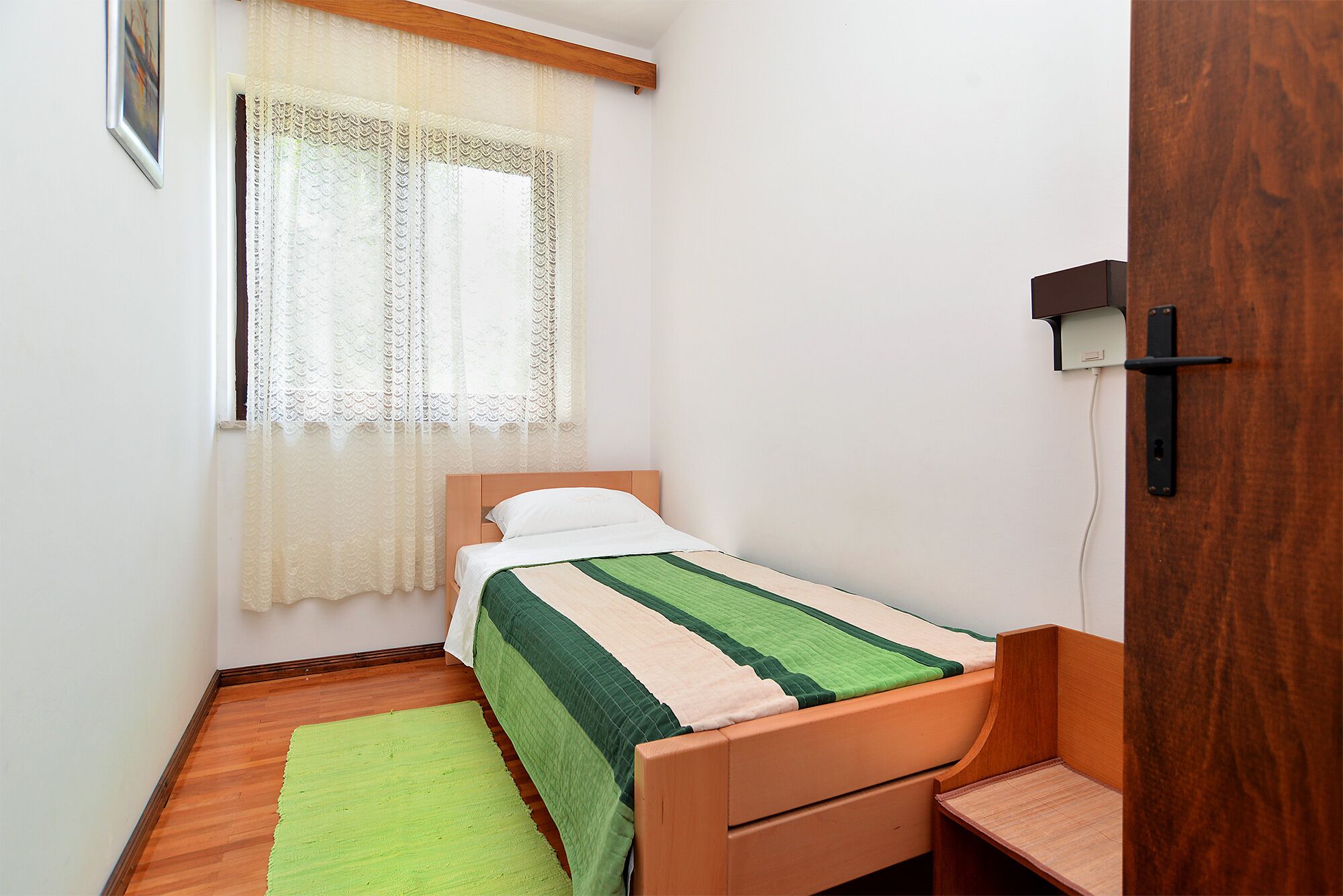 Een kleine, verzorgde slaapkamer met een eenpersoonsbed, een houten nachtkastje, een groen kleed en een raam met kantgordijnen.