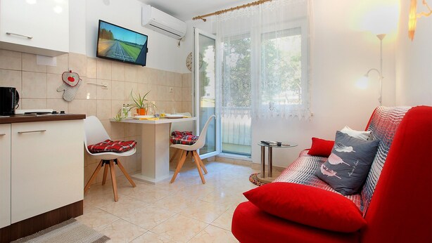 Un salon moderne avec un canapé rouge, un coin repas avec une petite table et une cuisine dotée de meubles blancs et d’un téléviseur.