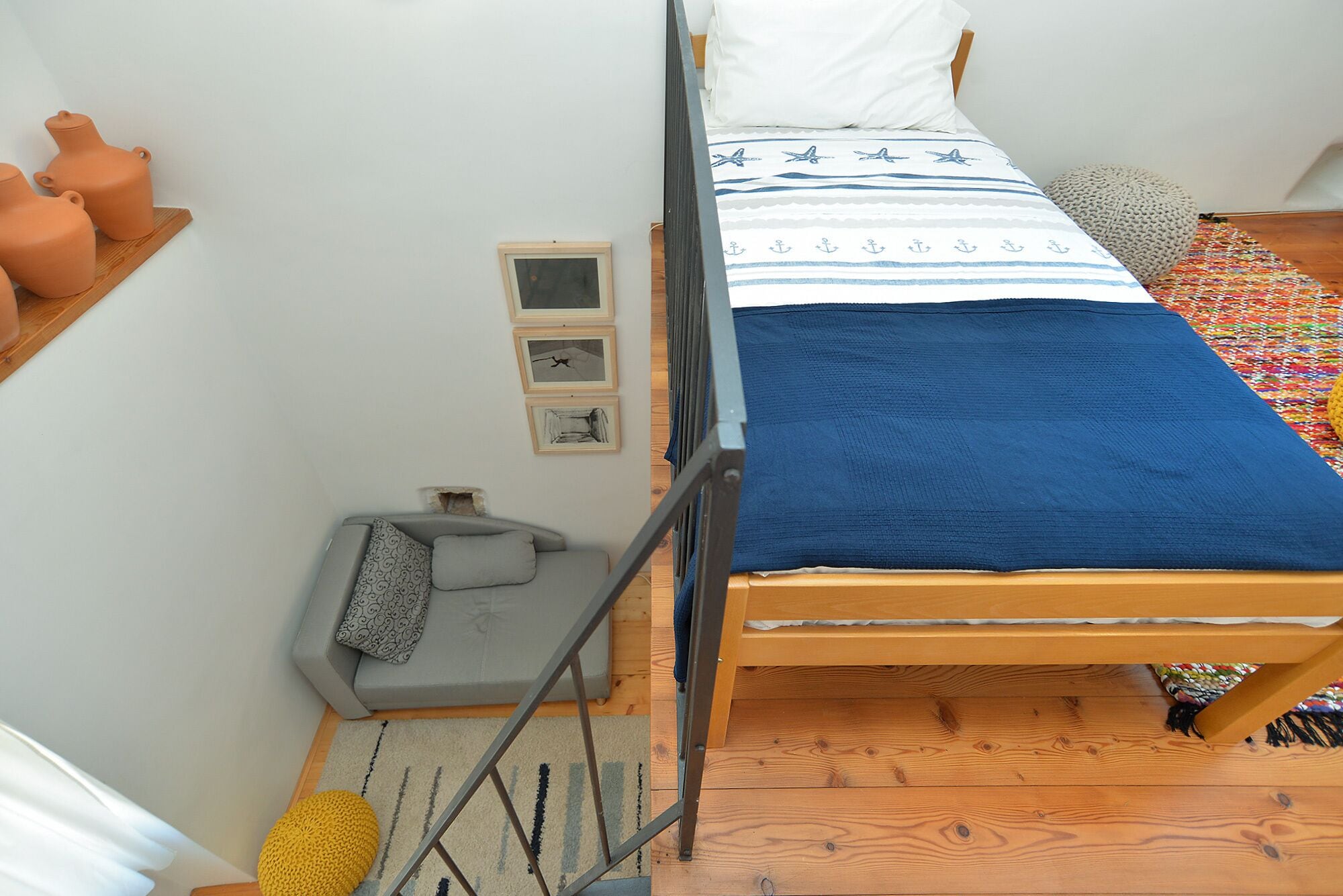 Un dormitorio pequeño y acogedor con una cama, suelo de madera, un sofá gris, un tapete pequeño y una estantería con objetos decorativos.