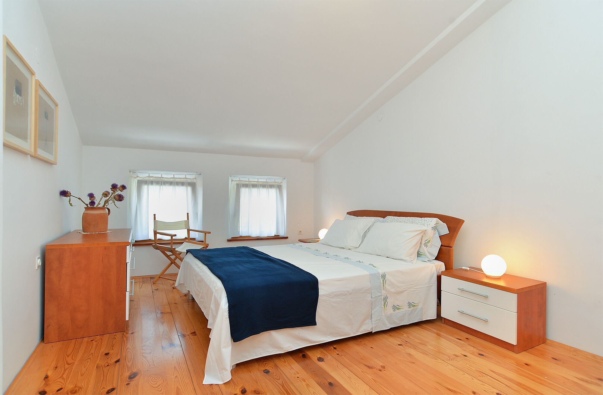 A bedroom with a bed, bedside tables, a dresser, a chair, and two windows with curtains.