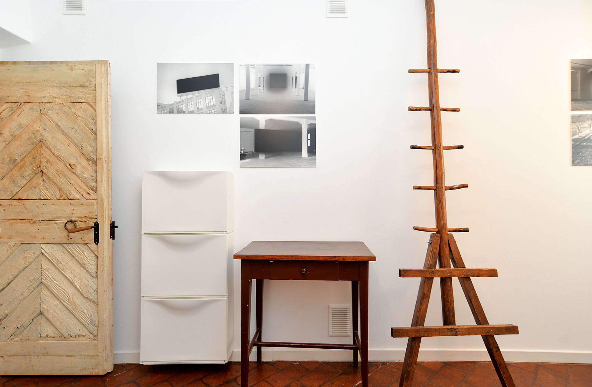 A room with a wooden door, a white refrigerator, a wooden table, and a wooden easel against a white wall with three black-and-white photographs.