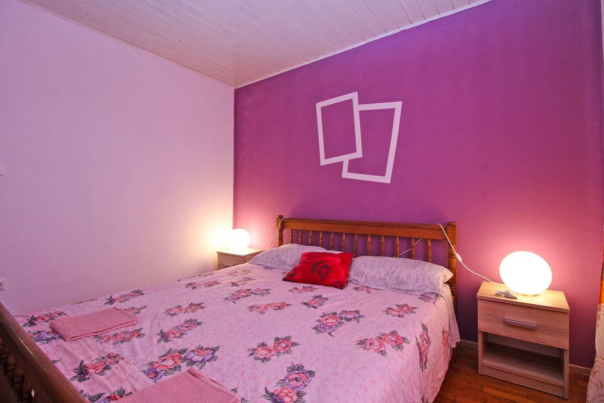 A bedroom with a wooden bed, floral bedding, two bedside lamps, and a purple wall with a white geometric design.