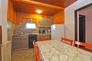 A compact kitchen with wooden cabinets, a stove, a refrigerator, and a dining table with chairs. - Apartment 1954 (Fazana)
