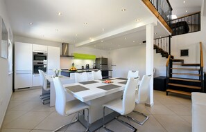 Een moderne keuken met witte kasten, een eethoek met een witte tafel en stoelen, en een trap met houten treden en zwarte leuningen.