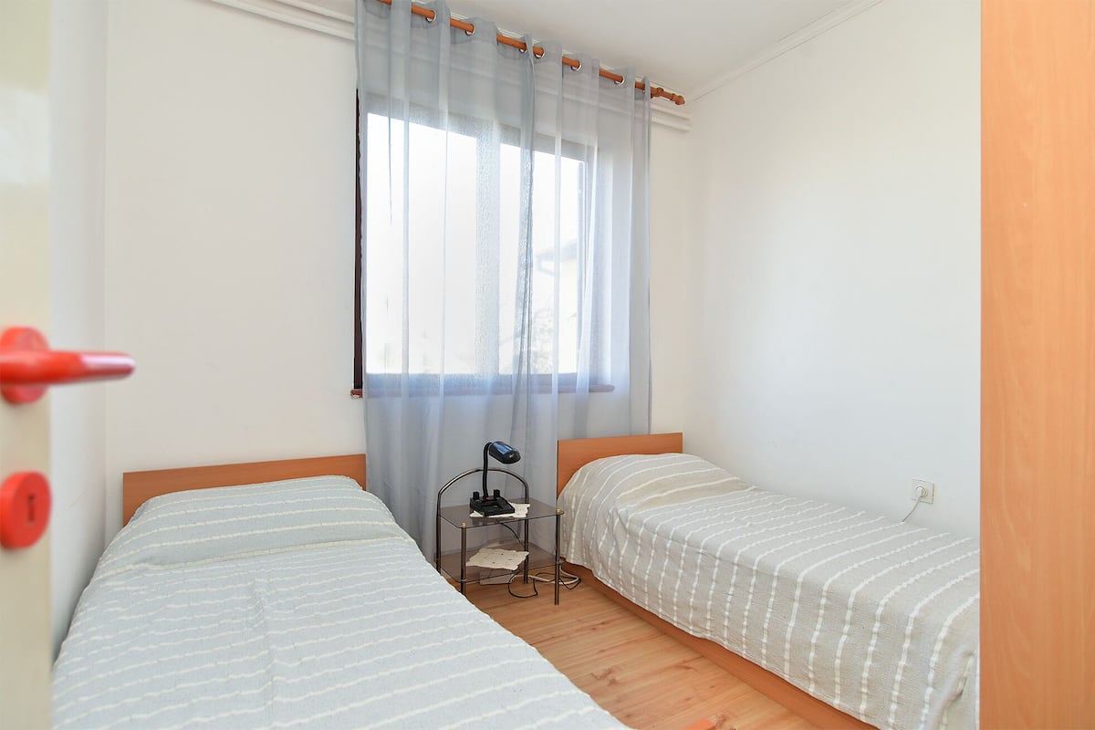A small, clean room with two single beds, a window with sheer curtains, and a bedside table with a lamp.