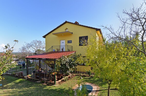 A yellow house with a red roof, a balcony, and a garden with a small tree and a blue chair. - Apartment 1956 (Pula)